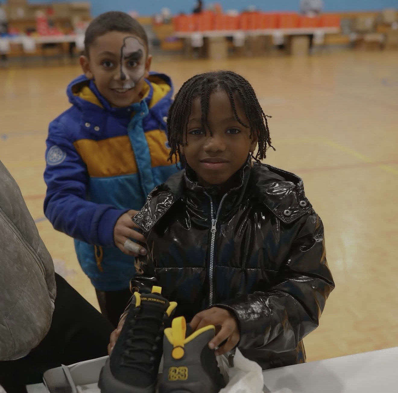 A boy holding up a new Air Jordan sneaker at a giveaway in a school gym, with another child in face paint standing behind him.