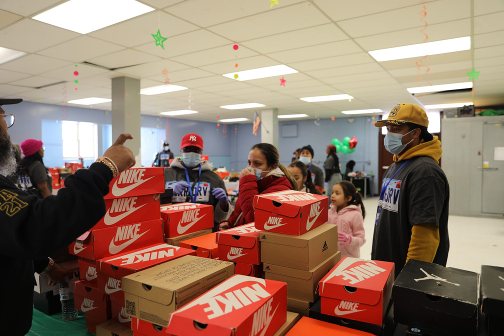 Stacks of Nike shoeboxes on a table at a giveaway, with volunteers and families gathered around them in a community room hung with holiday decorations.