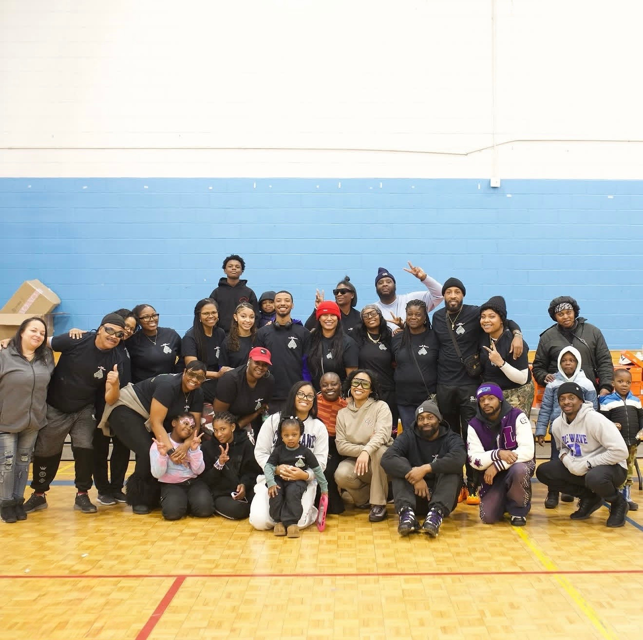 Volunteers and families at a past sneaker drive, gathered for a group photo in a school gym. Most are wearing matching black Ladi Luck t-shirts, with kids kneeling in front and donation boxes stacked against the wall.